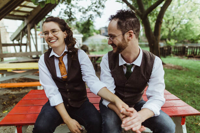 A picture of Alison and Jake, both young white people, sitting on a red picnic table. They are holding hands. Jake is looking at Alison, who is grinning and laughing at something he just said. Both are wearing jeans, white shirts, brown vests, and ties.
