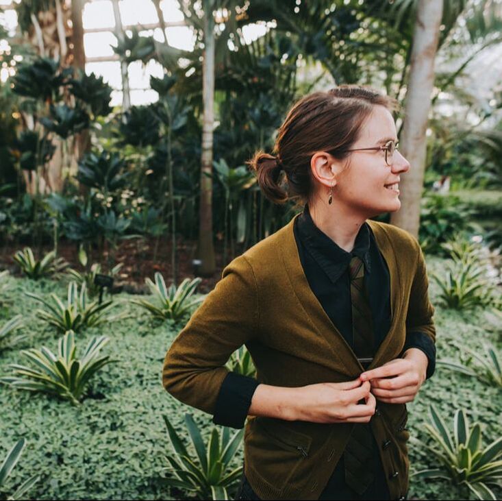 Picture of Alison, a young-ish white person with brown hair pulled back. She is wearing a black button down, green cardigan, and green tie (with a tie clip sensuously peeking out). She is smiling and looking to one side. She's in a conservatory, with greenery behind her.