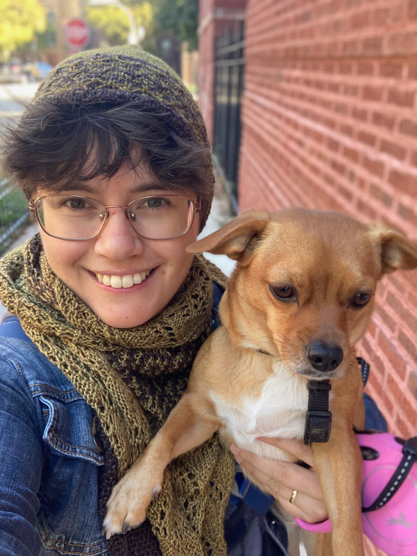 A picture of Alison, a young-ish white woman, holding Jo, a small brown dog. Alison is wearing a hand-knit hat and scarf, both green and brown. She is smiling at the camera.